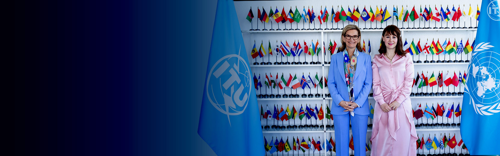 Assistant secretary Arielle Roth and Doreen Bogdan-Martin, Secretary-General of the International Telecommunication Union in front of shelves filled with small international flags, with large blue International Telecommunication Union flag on the left side